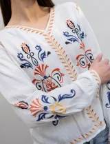 Blusa blanca de rayón con escote en V y mangas largas. Presenta bordados florales en tonos naranja, azul y negro en la parte delantera y en las mangas.
