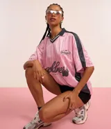 Remera rosa con cuello polo gris y rayas verticales. Estampado frontal con la palabra "gobline" en letras cursivas y la frase "in my best era". Diseño oversized con mangas cortas y recortes laterales en color negro.