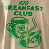 Remera oversize color beige con estampado en la espalda de una taza de café con cara sonriente, un cenicero con un cigarrillo y la frase "420 Breakfast Club. Wake and bake everyday".