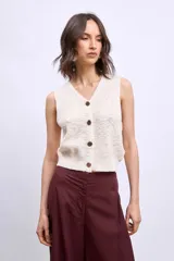 Chaleco de tejido de punto color crudo, sin mangas, con escote en V y cierre frontal de botones marrones.