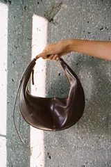 Bolso de mano ovalado color marrón oscuro, con asa de hombro metálica dorada y cierre superior con cremallera.