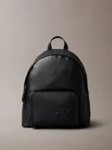Mochila negra de diseño moderno con acabado tipo piel sintética. Presenta un asa de mano superior, correas ajustables para el hombro, compartimento principal con cremallera y un bolsillo exterior frontal con el monograma 'CK' grabado en relieve.