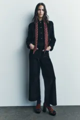 Pantalón ancho de pana color negro, de tiro medio, con trabillas en la cintura, bolsillos delanteros y cierre frontal con cremallera, botón y gancho metálico.