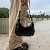 Cartera de hombro negra, estilo baguette, con acabado charol y textura de cocodrilo. Posee correa corta ajustable con detalle de nudo y herrajes dorados.