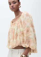 Blusa corta de gasa color crema con estampado floral en tonos durazno y verde. Tiene escote cuadrado, mangas largas abullonadas y espalda abierta.