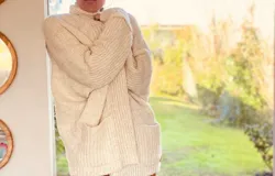 Cárdigan largo color crudo, de tejido de punto grueso con cuello alto, manga larga y bolsillos delanteros.