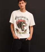 Remera color crema de corte recto, con cuello redondo y mangas cortas. Presenta un estampado estilo tatuaje con un felino en llamas y la frase "Redención es una virtud".