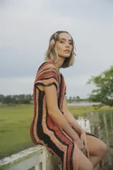 Vestido corto tejido a crochet con lurex, con escote en V y mangas cortas. Diseño a rayas horizontales en tonos naranja, marrón y negro, con flecos en el ruedo.