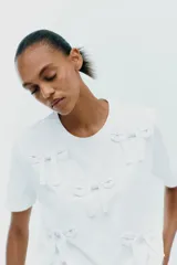 Camiseta blanca de algodón con cuello redondo, manga corta y apliques de lazos de raso blancos en el frente.