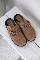 Mules destalonados de gamuza color camel, con tira ancha decorada con ojales metálicos y hebilla ajustable. Suela negra gruesa.