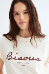 Remera color crema de corte holgado en punto de algodón. Modelo con cuello redondo y estampado frontal con la palabra "Bisous" en color borgoña y la frase "Mon Paris 1985" en negro.