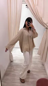 Camisa color beige claro con textura brillante, cuello clásico, cierre frontal con botones y mangas largas con puños abotonados.