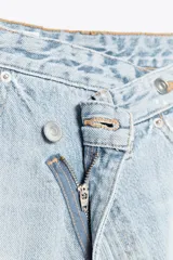 Pantalón de jean celeste de tiro alto, corte wide leg y largo hasta el tobillo. Presenta un diseño asimétrico en la cintura con cierre frontal cruzado con cremallera y botones metálicos.