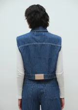 Chaleco crop de jean azul con rayas verticales blancas, cuello clásico, cierre con botones y bolsillos frontales con solapa. El corte es recto y llega hasta la cintura.