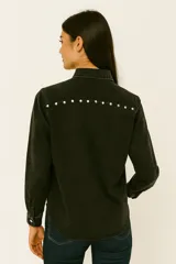 Camisa de jean negra con cuello clásico, cierre frontal con botones a presión, dos bolsillos con solapa en el pecho y tachas plateadas decorativas en cuello, hombros y bolsillos.