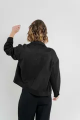 Chaqueta color beige de corte recto, estilo camisa, confeccionada en gamuza con elastano. Presenta cuello camisero, cierre frontal con botones metálicos, bolsillos grandes en el frente y charreteras en los hombros.