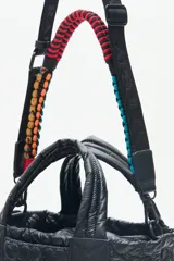 Cartera tote azul de nylon matelassé con diseño de corazones. Tiene manijas cortas y dos correas desmontables, una de cuero sintético y otra de tela trenzada multicolor. Cuenta con cierre superior y dos bolsillos internos.