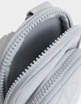 Portacelular gris acolchado de tela deportiva con bolsillo frontal con cierre y puller de goma. Tiene correa regulable y desmontable y cierre superior.