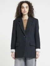 Blazer de corte recto, color negro o azul muy oscuro, con finas rayas verticales en tono claro (posiblemente marrón o beige). Presenta solapas de muesca, cierre frontal de dos botones y bolsillos de parche en la parte inferior. Se lleva con una camisa blanca a rayas finas asomando por las mangas.