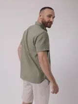 Camisa de hombre color verde oliva, de manga corta y corte clásico. Confeccionada en lino y poliéster, con cuello abotonado y bolsillo en el pecho.