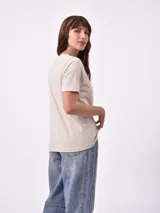 Remera blanca de algodón con cuello redondo y manga corta. Presenta un pequeño bordado en el pecho izquierdo con una figura que parece ser una persona acostada en una tabla de surf o flotador.