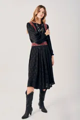 Vestido largo negro con estampado de hojas en relieve, mangas largas abullonadas y falda plisada. Presenta detalles bordados en rojo, blanco y azul en los hombros y la cintura.