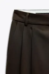 Pantalón marrón oscuro de tiro alto con trabillas, doble pliegue frontal, bolsillos laterales y falsos bolsillos de vivo en la espalda. Cierre frontal con cremallera y botón.