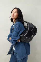Cartera negra tipo hobo de cuero sintético texturizado con apliques de tachas metálicas plateadas en forma de flor. Tiene asa corta y cierre superior con cremallera.