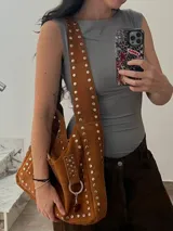 Cartera bandolera marrón de cuero con remaches metálicos plateados. Tiene correa ancha regulable, bolsillo externo y argolla para colgar accesorios.