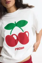 Remera blanca de algodón de corte amplio con cuello redondo y mangas cortas. Estampado frontal de dos cerezas rojas con hojas verdes y el logo de la marca Pacha en rojo.