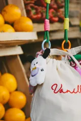 Llavero con mini peluche de perro blanco con orejas negras, babero lila y moño amarillo. Incluye correa de goma elástica en tonos rosa y celeste, dije de perla y mosquetón metálico rosa.