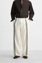 Pantalón relaxed fit confeccionado en tejido de lino color crudo. Cintura elástica ajustable con cordón, bolsillos laterales y traseros. Perneras con aplicación de bandas verticales negras a contraste en delantero.