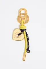 Llavero marca Bimba y Lola con mosquetón y anilla dorados. Presenta un dije metálico en forma de corazón con logo grabado, acompañado de un cordón de cuero amarillo y un cordón trenzado en azul marino y amarillo con nudos decorativos.