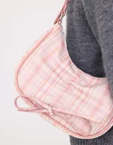 Bandolera de cuerina estampada con diseño escocés en tonos rosa y blanco. Presenta frunce y lazo frontal con moño, manija superior regulable con hebilla y cierre superior.