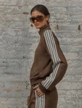 Conjunto deportivo marrón de dos piezas: campera con cremallera, cuello alto, mangas largas con puño y detalles de rayas blancas en mangas y laterales del pantalón. Pantalón recto de cintura alta con cordón ajustable.
