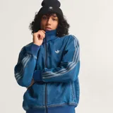 Campera deportiva Adidas Originals modelo Firebird, confeccionada en denim azul. Presenta cuello alto, cierre frontal, puños y cintura de tejido acanalado azul oscuro, y las tres franjas características de Adidas en las mangas en tono más claro. Incluye el logo del trébol bordado en blanco en el pecho.