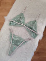 Conjunto de lencería de encaje floral color verde menta, con detalles y tiras ajustables en tono beige/tostado. Incluye un corpiño triangular sin aro y una bombacha tipo colaless o tanga.