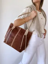 Bolso tipo tote de cuero marrón con costuras blancas a la vista y tiras decorativas. Asas de cuero color crema con detalle de trenzado. Forrada en tela.