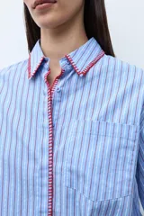 Camisa de manga larga, corte oversize, color celeste con rayas verticales blancas. Presenta un detalle de bordado en color rojo/fucsia a lo largo del cuello y la abertura frontal.