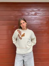 Sweater color crema tejido en lana merino, con apliques de cuero marrón y tejido trenzado en el frente. Presenta costuras a la vista en el ruedo y los puños.