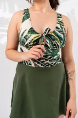 Vestido de baño enterizo con estampado de hojas de palmera en tonos verde, blanco y amarillo. Presenta escote redondo con nudo y abertura en el centro del busto, y falda corta verde oliva.