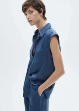 Camisa azul sin mangas de tejido satinado con cuello camisero, cierre abotonado y bolsillo de parche en el pecho.