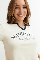 Remera corta color crema con cuello redondo y ribete azul marino. Estampado frontal con la inscripción "Manhattan New York City" en azul.