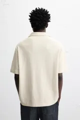 Camisa de manga corta color crudo, con cuello bowling, corte relaxed fit y cierre frontal con botones. Presenta apliques de parches con texturas combinadas a contraste en el delantero y aberturas laterales en el bajo.