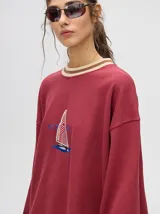 Buzo blanco de corte redondo (crew neck) con puños ajustados que tienen rayas rojas y blancas. Presenta un estampado central de un velero navegando con texto alrededor.