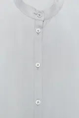 Camisa celeste fluida confeccionada en hilatura con mezcla de lyocell. Cuello redondo, manga larga con puños y bajo asimétrico con aberturas laterales. Cierre frontal con botones.