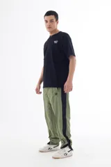 Pantalón de jogging verde oliva con banda lateral negra, confeccionado en nylon con forro de red, corte recto y cierres en los tobillos.