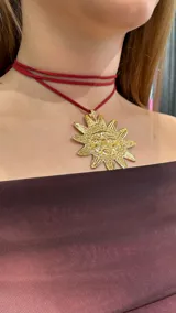 Gargantilla tipo choker con doble tira de gamuza roja y dije dorado con forma de sol con rostro.