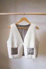 Chaleco marrón de cuero con interior de corderito color crema, cierre con gancho metálico y bolsillos frontales de corderito.