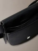 Cartera bandolera negra de cuero sintético con textura granulada, forma de media luna y correa fina ajustable. Cierre con solapa magnética y logo de Calvin Klein grabado en el frente.